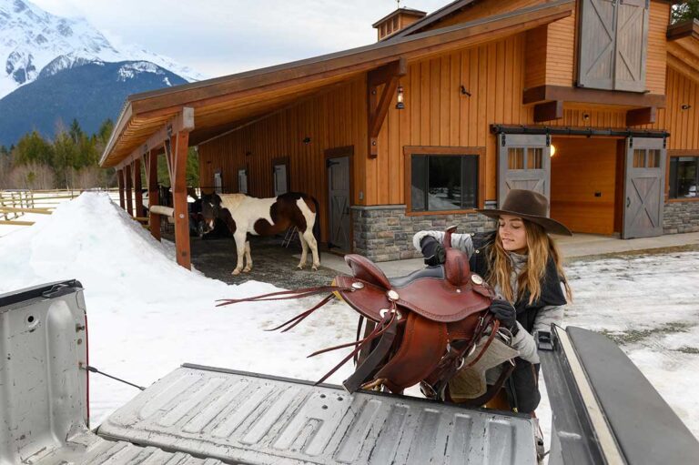a woman lifts her western saddle into the back of her pickup truck on a snowy day, with her paint horse and barn in the background