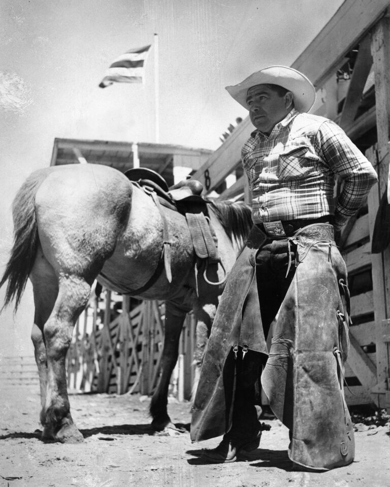 Black and white photo of Ed Yanez standing in front of a saddle horse.