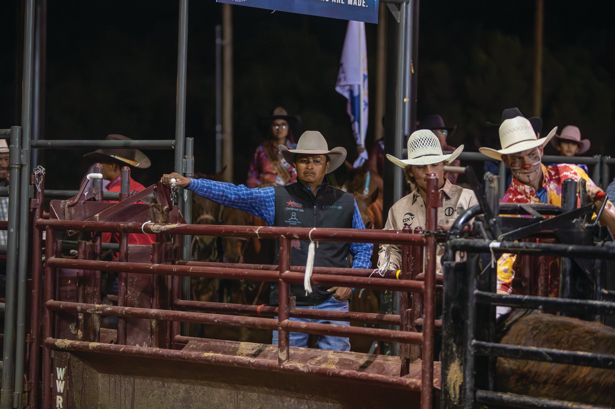 Contestants and a rodeo clown gather behind the chutes at night at the Home of the Navajo PRCA Rodeo