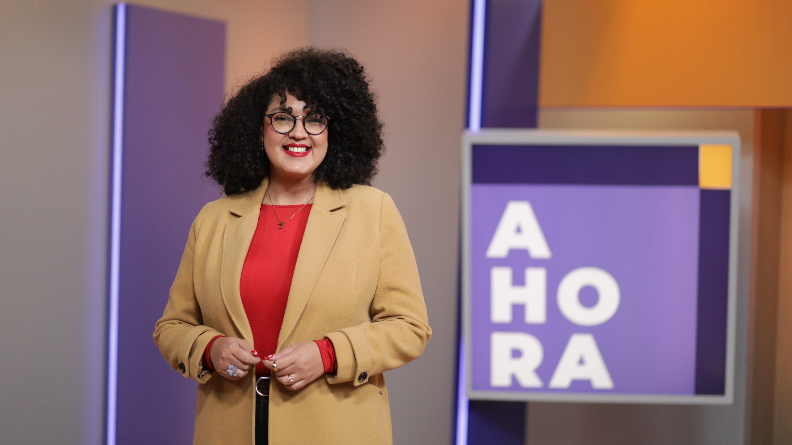 Jenny Bastidas, periodista cultural de AHORA, vestida de blazer beige y camisa roja, sonriendo en el set de AHORA.