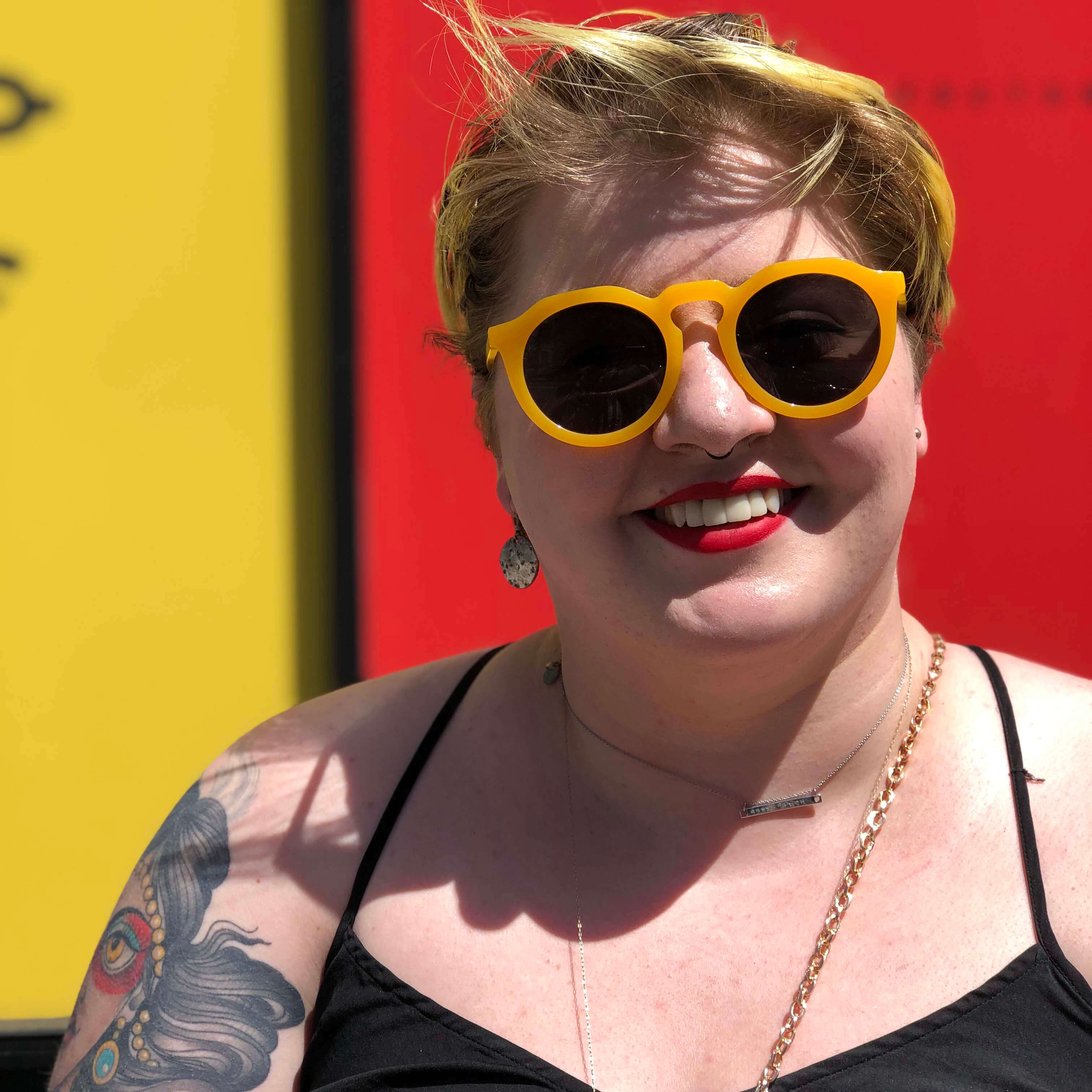 nikki bosso in yellow sunglasses and black dress in front of red wall