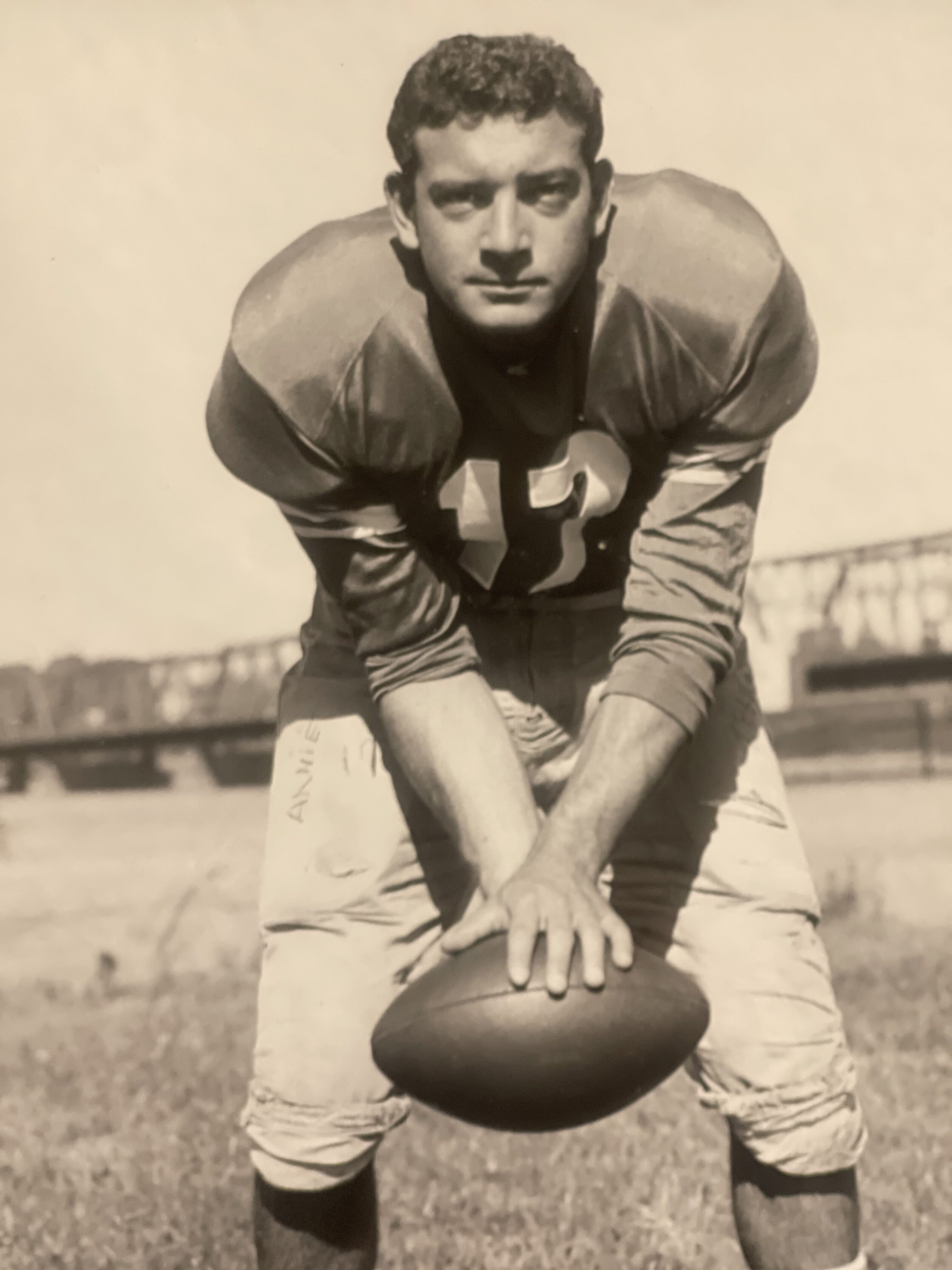 A football player in an old-fashioned uniform, wearing number 12, poses outdoors holding a football with both hands, with a bridge visible in the background.