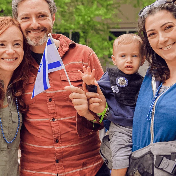 Two women and a man smiling for a photo. Two are waving Israeli flags and the other is holding a toddler boy.