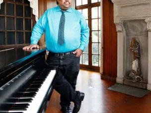 A man in a turquoise shirt and patterned tie stands by a grand piano in a wood-paneled room with large windows and a decorative fireplace.