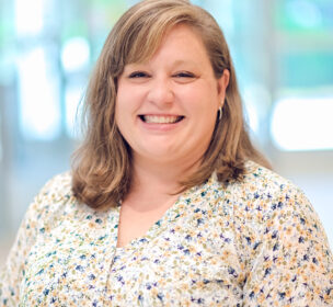A woman with shoulder-length light brown hair smiles at the camera, wearing a light floral blouse, standing indoors in a brightly lit setting.
