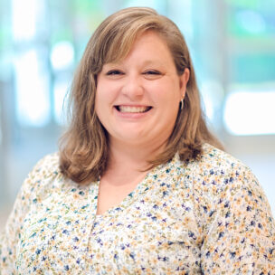 A woman with shoulder-length light brown hair smiles at the camera, wearing a light floral blouse, standing indoors in a brightly lit setting.