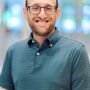 A man with glasses and a short beard, wearing a green striped polo shirt, smiles while standing indoors with a blurred background.