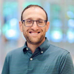 A man with glasses and a short beard, wearing a green striped polo shirt, smiles while standing indoors with a blurred background.