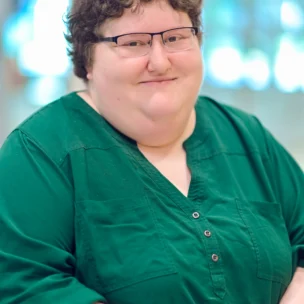 A person with short curly hair and glasses wearing a green shirt stands indoors with hands clasped, smiling at the camera.