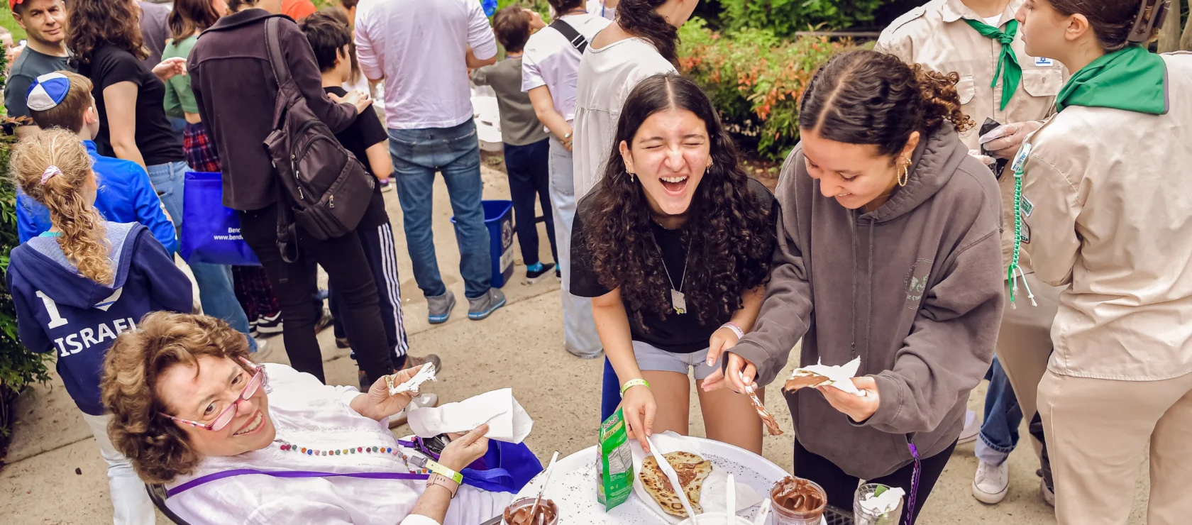 Two girls outside laughing together while making food. An older woman sits near them smiling at the camera.