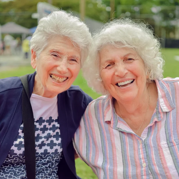Two elderly women smiling and sitting next to each other outdoors, with a grassy area and soccer goal visible in the background.