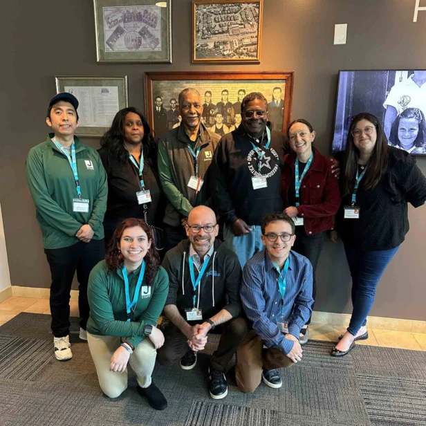 A group of nine people standing and kneeling indoors, wearing ID badges and casual clothing, posing in front of framed pictures on the wall.