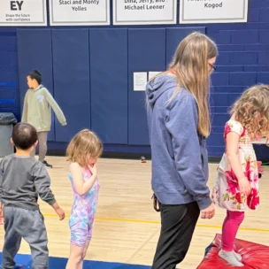 Four young children and one adult stand and walk on blue mats in a gymnasium with a blue wall and posters in the background.