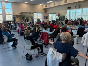 A group of adults sit in chairs in a fitness studio, participating in an exercise class using resistance bands, with large mirrors and windows in the background.