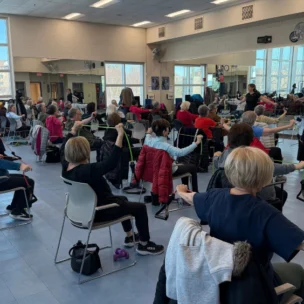 A group of adults sit in chairs in a fitness studio, participating in an exercise class using resistance bands, with large mirrors and windows in the background.