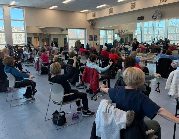 A group of adults sit in chairs in a fitness studio, participating in an exercise class using resistance bands, with large mirrors and windows in the background.