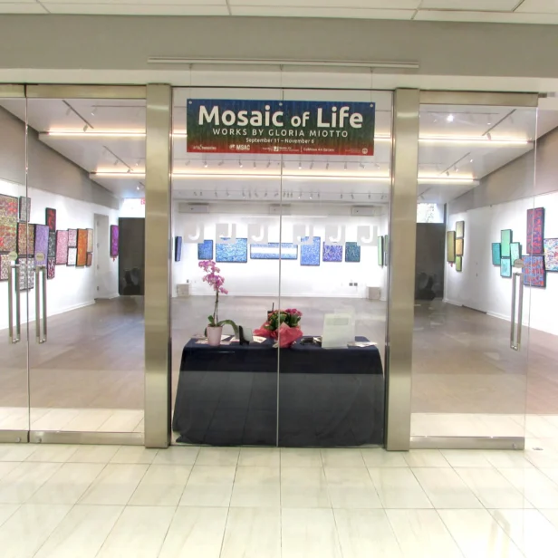 An art gallery with glass doors displays colorful framed artworks on white walls. A banner reads "Mosaic of Life." A black table holds pink flowers in the foreground.
