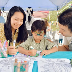 A child colors a picture at a table, surrounded by art supplies, with a woman and man attentively watching under a canopy.