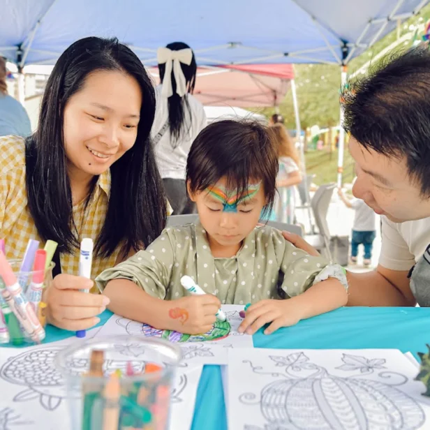 A child colors a picture at a table, surrounded by art supplies, with a woman and man attentively watching under a canopy.