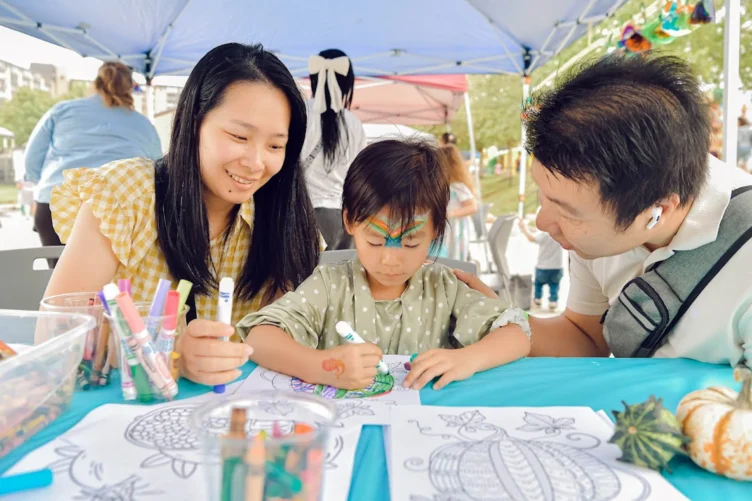 A child colors a picture at a table, surrounded by art supplies, with a woman and man attentively watching under a canopy.