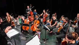 Orchestra performing on stage with various string instruments; musicians surrounded by children observing closely.