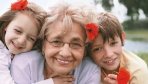 An elderly woman with glasses, smiling between a young girl and boy. Each has a red flower in their hair. Outdoors with greenery and water in the background.