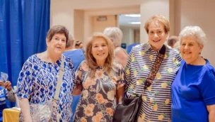 Four smiling women stand together indoors, wearing colorful tops, with a blue curtain and a table in the background.