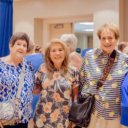 Four smiling women stand together indoors, wearing colorful tops, with a blue curtain and a table in the background.