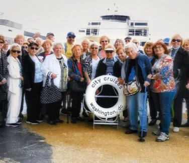 A group of people, mostly older adults, stand together on a dock in front of a "City Cruises Washington DC" lifebuoy, with parts of a boat visible in the background.