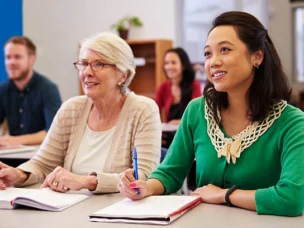Two women seated at a desk in a classroom, taking notes and listening attentively, with other students visible in the background.
