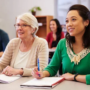 Two women seated at a desk in a classroom, taking notes and listening attentively, with other students visible in the background.