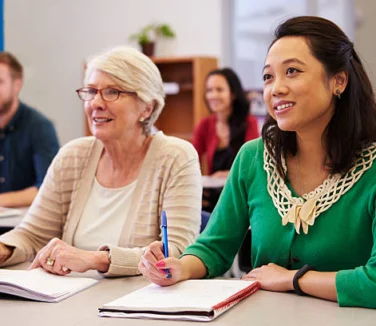 Two women seated at a desk in a classroom, taking notes and listening attentively, with other students visible in the background.