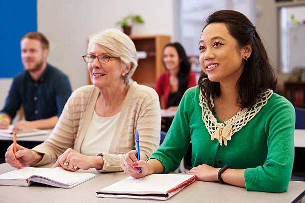 Two women seated at a desk in a classroom, taking notes and listening attentively, with other students visible in the background.