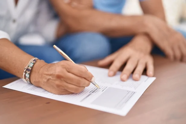 Close-up of a person signing a document on a wooden table, with another person's hand resting nearby.