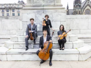 Four musicians pose outdoors on marble steps with their string instruments: two violins, a viola, and a cello. Stone buildings and a church are visible in the background.