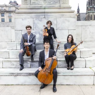Four musicians pose outdoors on marble steps with their string instruments: two violins, a viola, and a cello. Stone buildings and a church are visible in the background.