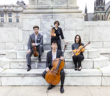 Four musicians pose outdoors on marble steps with their string instruments: two violins, a viola, and a cello. Stone buildings and a church are visible in the background.