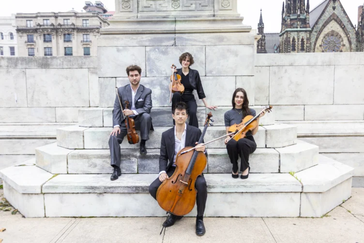 Four musicians pose outdoors on marble steps with their string instruments: two violins, a viola, and a cello. Stone buildings and a church are visible in the background.