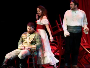 Three actors perform on stage; one man sits in a military uniform, a woman in a white dress stands behind him, and another man in a white shirt stands to the right with chairs in the background.