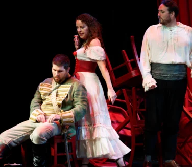 Three actors perform on stage; one man sits in a military uniform, a woman in a white dress stands behind him, and another man in a white shirt stands to the right with chairs in the background.