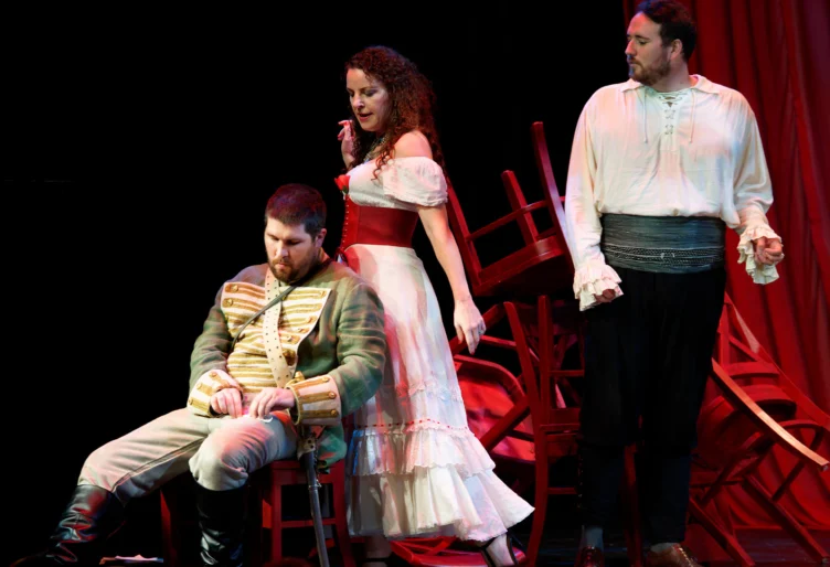 Three actors perform on stage; one man sits in a military uniform, a woman in a white dress stands behind him, and another man in a white shirt stands to the right with chairs in the background.