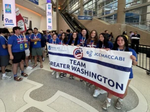 A group of teenagers in blue shirts hold a "Team Greater Washington" banner inside a large indoor venue with signs and monitors in the background.