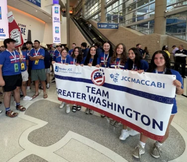 A group of teenagers in blue shirts hold a "Team Greater Washington" banner inside a large indoor venue with signs and monitors in the background.