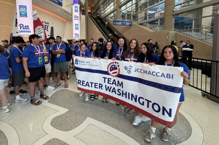 A group of teenagers in blue shirts hold a "Team Greater Washington" banner inside a large indoor venue with signs and monitors in the background.