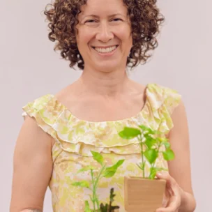 A woman with curly hair in a yellow floral top smiles while holding two small potted plants against a plain background.