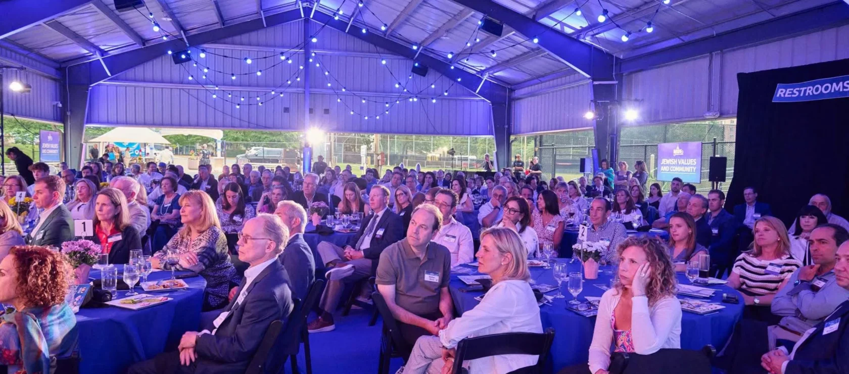 A large group of people seated at tables under a blue-lit event tent, attentively listening to a presentation.