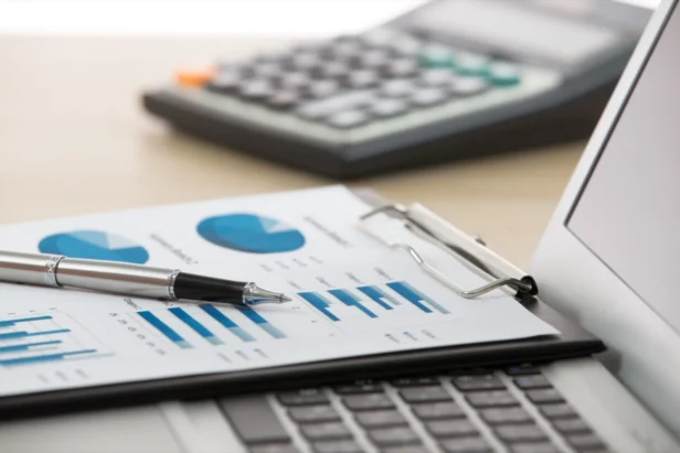 A pen rests on financial charts with blue graphs on paper, next to a laptop keyboard and a blurred calculator in the background.