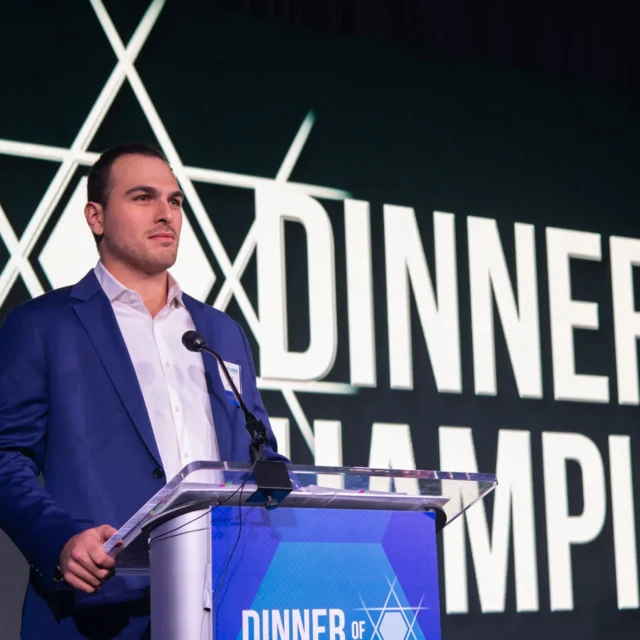 Man in a blue suit speaks at a clear podium with a microphone, in front of a large "Dinner of Champions" sign.