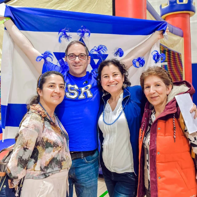Four people smiling, standing together indoors. One person holds a flag and wears festive accessories. There's a bouncy castle in the background.