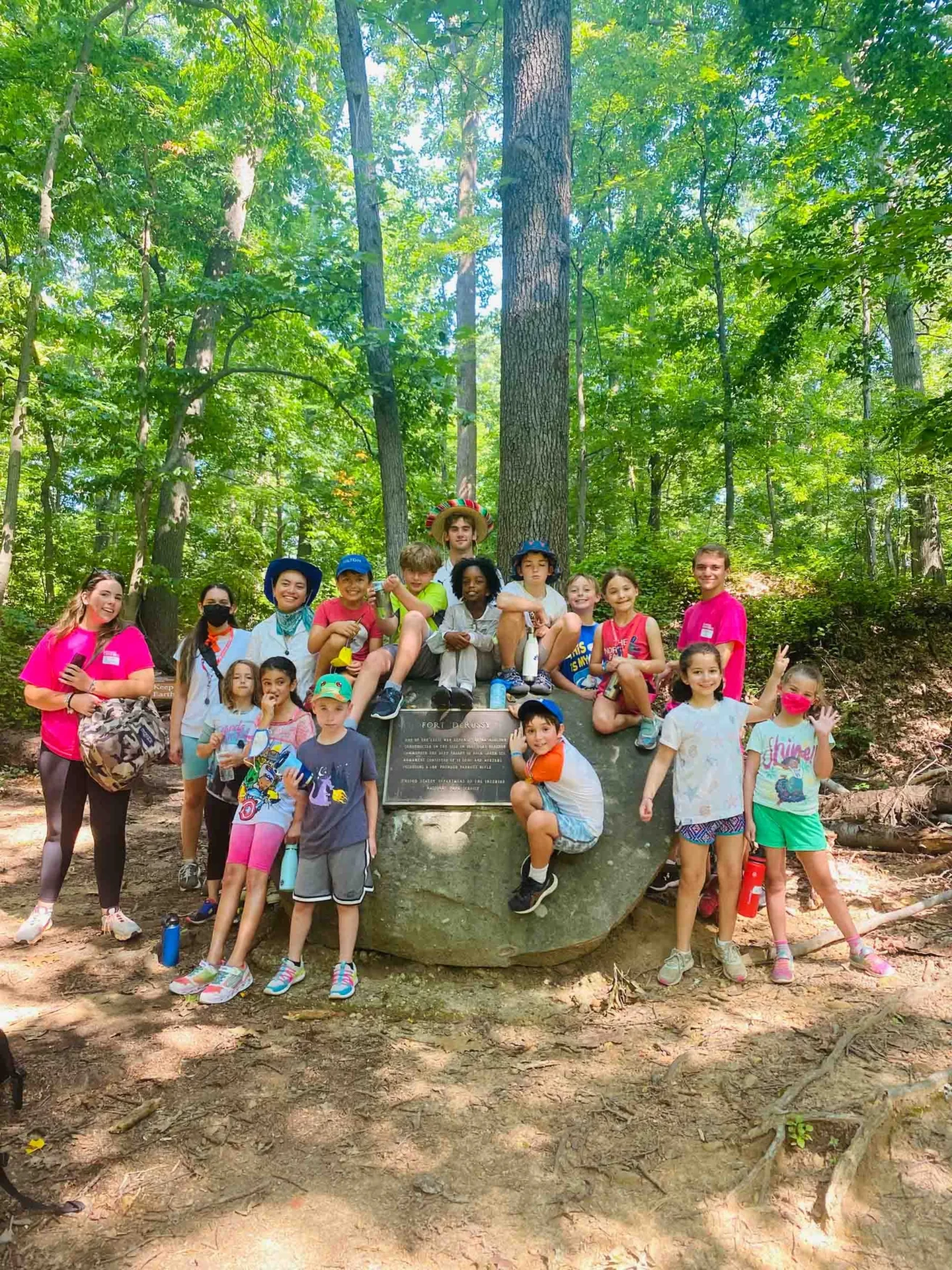 A group of children and an adult pose in a wooded area, sitting and standing around a large rock with a plaque. Some children wear hats and colorful clothing.